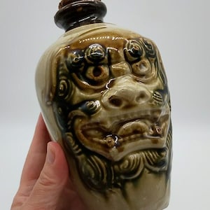 May include: A ceramic bottle with a glossy beige and dark brown glaze, featuring a prominent relief sculpture of a lion-like face on the front. The bottle has a narrow neck topped with a round cork stopper, showcasing intricate moulded details.