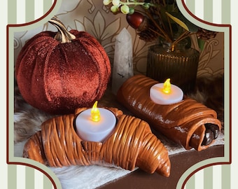 May include: A seasonal display with a velvet pumpkin and ceramic candle holders shaped like a croissant and a pain au chocolat. Each pastry-shaped holder contains a battery-operated candle with a flickering yellow light.