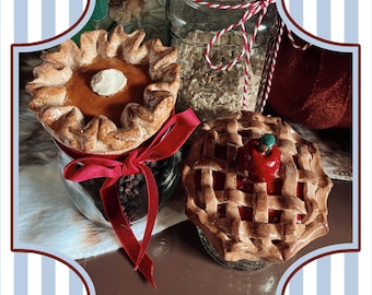 May include: Two miniature pie ornaments, one pumpkin and one apple, sit in glass jars. The pumpkin pie has a dollop of white cream. The apple pie has a lattice crust and a red apple on top. A red ribbon adorns the pumpkin pie jar.