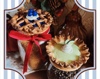 May include: Two decorative pie-shaped jar toppers. One is a blueberry pie with a lattice crust, the other is a key lime pie with whipped cream and lime slices. Both are set on glass jars with decorative fillings and a red ribbon.