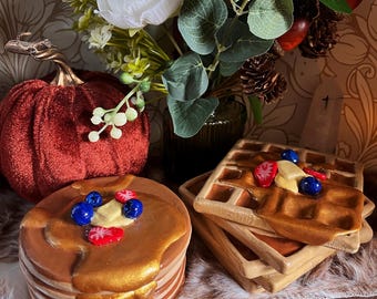 May include: A stack of brown pancakes with golden syrup, blueberries, and strawberries. Beside it are several brown waffles with similar toppings. A red velvet pumpkin and a bouquet of flowers are in the background.