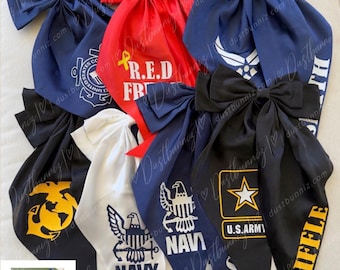 May include: A collection of large fabric hair bows featuring various military branch emblems and text including R.E.D. FRIDAY, NAVY, U.S. ARMY, and RIFLE. The bows are in red, white, navy blue, and black, with a small inset showing the back of a bow.