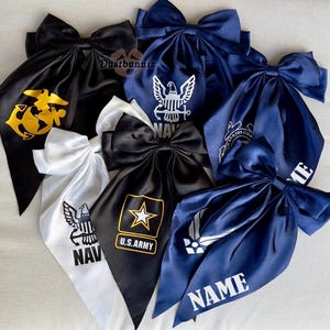May include: A collection of satin hair bows in black, white, and navy blue. Each bow features a military emblem and text, including the U.S. Army star, Navy eagle, and Air Force logo. The bows are tied with a large knot.