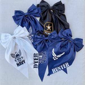May include: A collection of five large fabric hair bows in navy blue, black, and white. Each bow features military branch emblems and text including Navy, U.S. Army, Coast Guard, and Air Force, with personalized names like Dyer and Hunter printed.