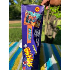 Puede incluir: Una caja de figura de acci&oacute;n de Spider-Man morada y azul. La caja presenta el logotipo de Spider-Man, arte de c&oacute;mic y texto que dice "10" de alto, totalmente posable, accesorio incluido". La caja es de la edici&oacute;n Deluxe.