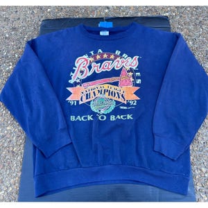 May include: A navy blue sweatshirt featuring the Atlanta Braves logo in pink, white, and red. The graphic includes the text "Braves," "National League Champions," and "Back to Back," with the years '91 and '92. A vintage sports apparel item.