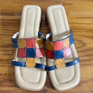 May include: A pair of slip-on sandals with a square toe and a low block heel. The upper features a woven, multi-colored leather strap design in red, yellow, and navy blue, secured with visible stitching. The insole is beige and stamped with the brand name DANELLE.