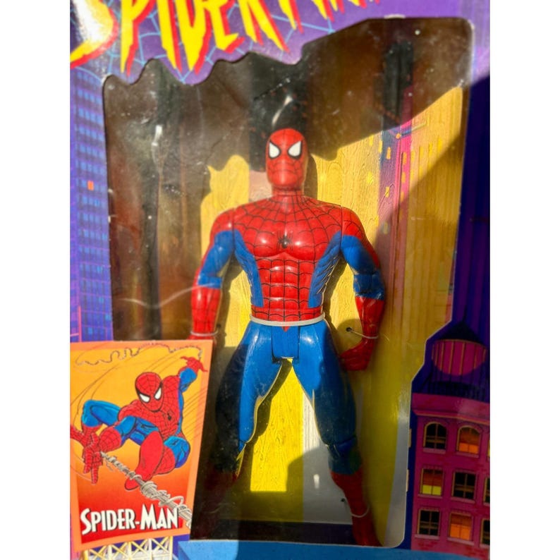 Puede incluir: Figura de acci&oacute;n de Spider-Man en su caja. La figura es roja y azul, con un dise&ntilde;o detallado. La caja presenta el logotipo de Spider-Man y una ilustraci&oacute;n de estilo c&oacute;mic del personaje. El fondo tiene un dise&ntilde;o de paisaje urbano.