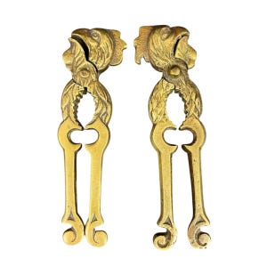 Op de afbeelding: Twee bijpassende notenkrakers van messing met een gedetailleerd ontwerp van een adelaarskop aan de bovenkant. De handvatten zijn gevormd met sierlijk, gebogen metaalwerk en textuurdetails, met een metallic gouden afwerking over de structuur.