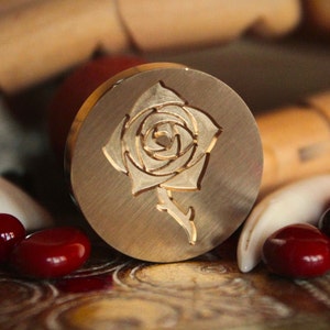 May include: A brass wax seal stamp with a detailed design of a rose. The rose is engraved in the center of the stamp and has a stem and leaves.