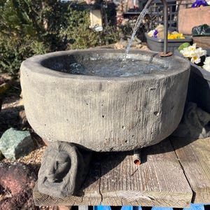 Peut inclure: Une fontaine en pierre grise patin&eacute;e avec de l'eau coulant dans un bassin rond. La fontaine repose sur une surface en bois, avec une figure en pierre sculpt&eacute;e &agrave; la base. Des fleurs blanches et jaunes sont dans un pot &agrave; proximit&eacute;.