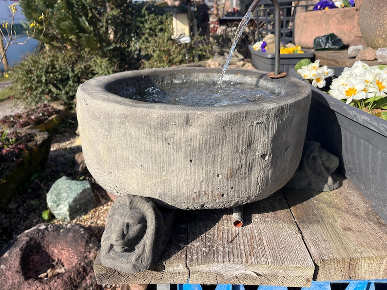 Peut inclure: Une fontaine en pierre grise patin&eacute;e avec de l'eau coulant dans un bassin rond. La fontaine repose sur une surface en bois, avec une figure en pierre sculpt&eacute;e &agrave; la base. Des fleurs blanches et jaunes sont dans un pot &agrave; proximit&eacute;.