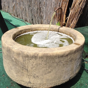 May include: A stone fountain with a spout pouring water into a basin. The water is green and bubbly. The fountain is surrounded by green grass.