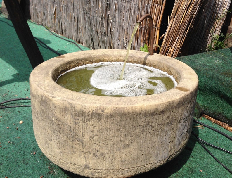 May include: A stone fountain with a spout pouring water into a basin. The water is green and bubbly. The fountain is surrounded by green grass.