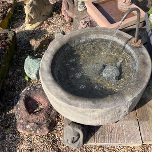 Peut inclure: Une fontaine en pierre grise avec de l'eau coulant d'un bec m&eacute;tallique dans un bassin rond. La fontaine est entour&eacute;e de roches, de plantes et d'une surface en bois. Des fleurs blanches et jaunes sont visibles dans une jardini&egrave;re voisine.