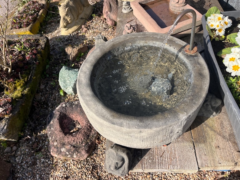 Peut inclure: Une fontaine en pierre grise avec de l'eau coulant d'un bec m&eacute;tallique dans un bassin rond. La fontaine est entour&eacute;e de roches, de plantes et d'une surface en bois. Des fleurs blanches et jaunes sont visibles dans une jardini&egrave;re voisine.