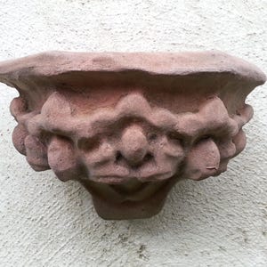 Może przedstawiać: Rzeźba z czerwono-brązowego kamienia przedstawiająca groteskową twarz z szerokimi ustami i okrągłym nosem. Rzeźba jest rzeźbiona z misternymi detalami i wydaje się być elementem dekoracyjnym budynku.