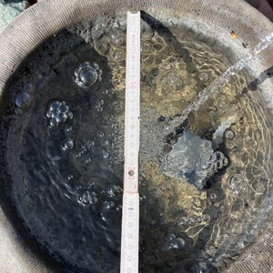 Peut inclure: Un abreuvoir rond en pierre grise rempli d'eau. Une r&egrave;gle en bois, gradu&eacute;e en centim&egrave;tres, est plac&eacute;e &agrave; l'int&eacute;rieur. Des bulles sont visibles dans l'eau, et un filet d'eau s'y d&eacute;verse. Le diam&egrave;tre du bain est d'environ 30 cm.