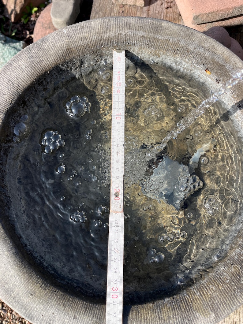 Peut inclure: Un abreuvoir rond en pierre grise rempli d'eau. Une r&egrave;gle en bois, gradu&eacute;e en centim&egrave;tres, est plac&eacute;e &agrave; l'int&eacute;rieur. Des bulles sont visibles dans l'eau, et un filet d'eau s'y d&eacute;verse. Le diam&egrave;tre du bain est d'environ 30 cm.