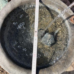 Peut inclure: Un abreuvoir en pierre rempli d'eau, avec une r&egrave;gle indiquant une profondeur d'environ 30 cm. L'eau coule dans le bain d'un bec m&eacute;tallique, cr&eacute;ant des bulles. L'abreuvoir a un design circulaire textur&eacute;.