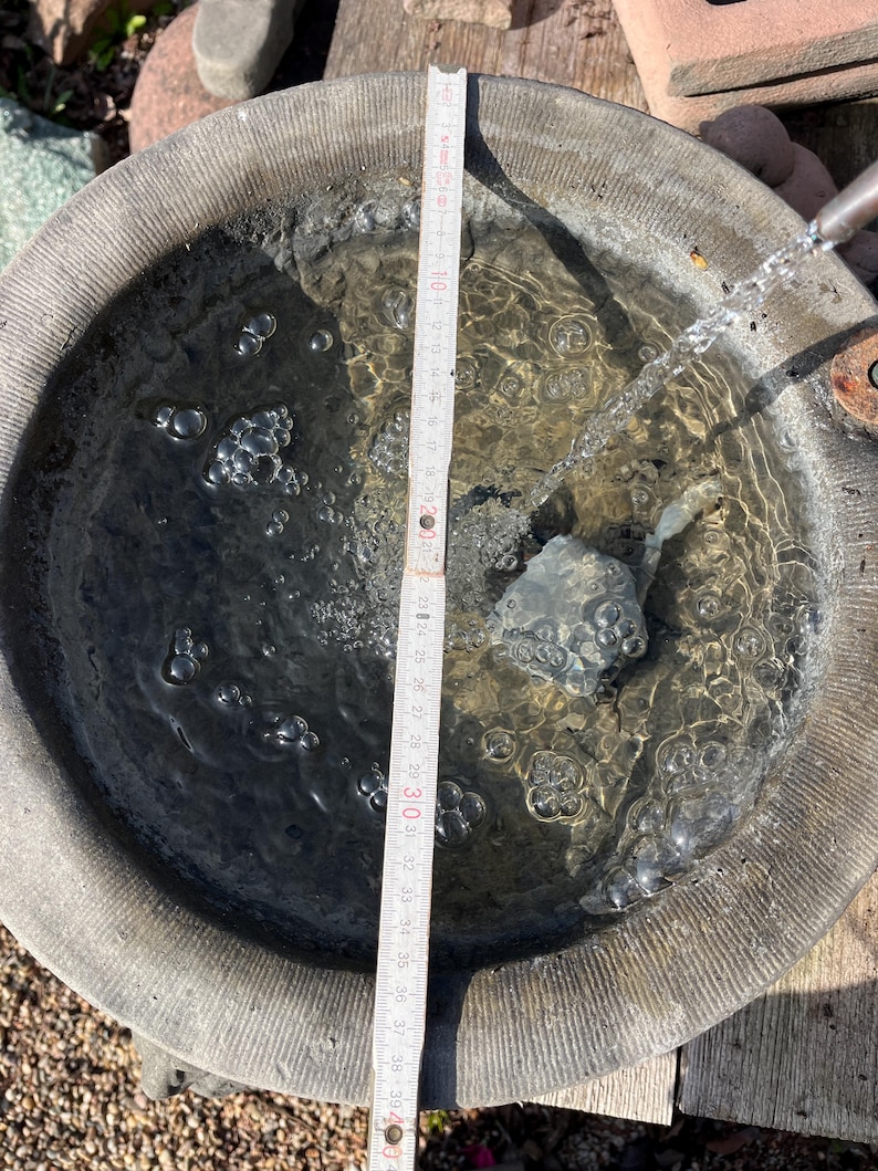 Peut inclure: Un abreuvoir en pierre rempli d'eau, avec une r&egrave;gle indiquant une profondeur d'environ 30 cm. L'eau coule dans le bain d'un bec m&eacute;tallique, cr&eacute;ant des bulles. L'abreuvoir a un design circulaire textur&eacute;.