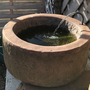 May include: A large, round, sandstone fountain with a water feature. The fountain is filled with water and has a small stream of water flowing from a pipe into the basin. The fountain is sitting on a stone base.