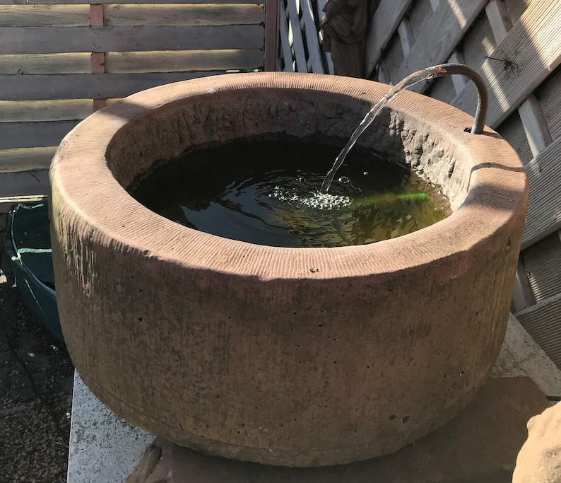May include: A large, round, sandstone fountain with a water feature. The fountain is filled with water and has a small stream of water flowing from a pipe into the basin. The fountain is sitting on a stone base.