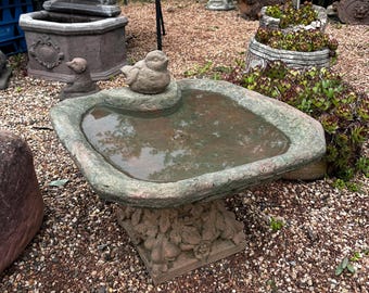 Op de afbeelding: Een stenen vogelbad met een klein vogelsculptuur erop. Het bad is gevuld met water en heeft een ruw, getextureerd oppervlak. Het vogelbad staat op een bed van kleine stenen.