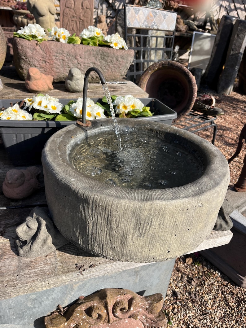 Peut inclure: Une fontaine en pierre grise avec de l'eau s'&eacute;coulant d'un bec incurv&eacute; dans un bassin rond. Des fleurs blanches et jaunes sont dans un bac en pierre derri&egrave;re la fontaine. La fontaine est sur une surface en bois.