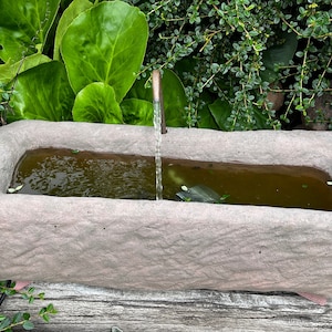 Könnte beinhalten: Ein grauer Steintrog mit einem kleinen Kupferrohr, aus dem Wasser in den Trog sprudelt. Der Trog ist mit Wasser gefüllt und von grünem Laub umgeben.