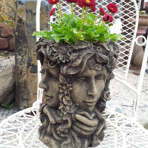 Può includere: Un vaso da fiori in cemento a forma di due volti con dettagli floreali, che contiene un vaso di fiori rossi e rosa.