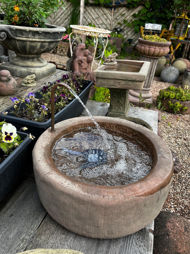 May include: A small, round, stone fountain with a water feature. The fountain is filled with water and has a small spout that is pouring water into the basin. The fountain is sitting on a wooden table.