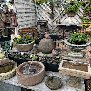 Peut inclure: Divers ornements de jardin en pierre, dont des abreuvoirs pour oiseaux, des jardini&egrave;res et des statues, sont expos&eacute;s sur une table en bois. Une petite fontaine est en activit&eacute;, et une statue d'oiseau est pos&eacute;e sur une sph&egrave;re en pierre. La sc&egrave;ne se d&eacute;roule dans un jardin avec une cl&ocirc;ture en bois.