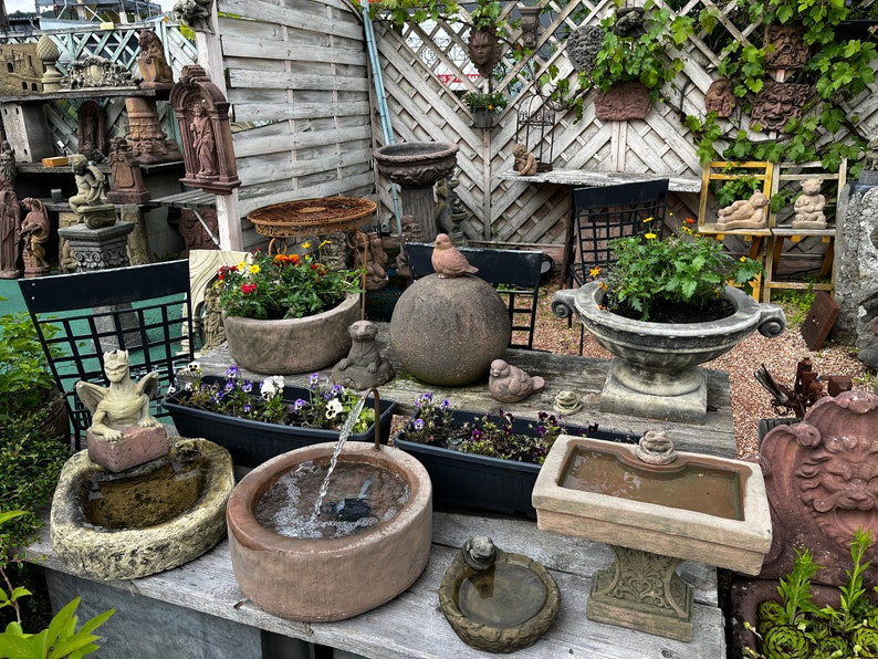 Peut inclure: Divers ornements de jardin en pierre, dont des abreuvoirs pour oiseaux, des jardini&egrave;res et des statues, sont expos&eacute;s sur une table en bois. Une petite fontaine est en activit&eacute;, et une statue d'oiseau est pos&eacute;e sur une sph&egrave;re en pierre. La sc&egrave;ne se d&eacute;roule dans un jardin avec une cl&ocirc;ture en bois.