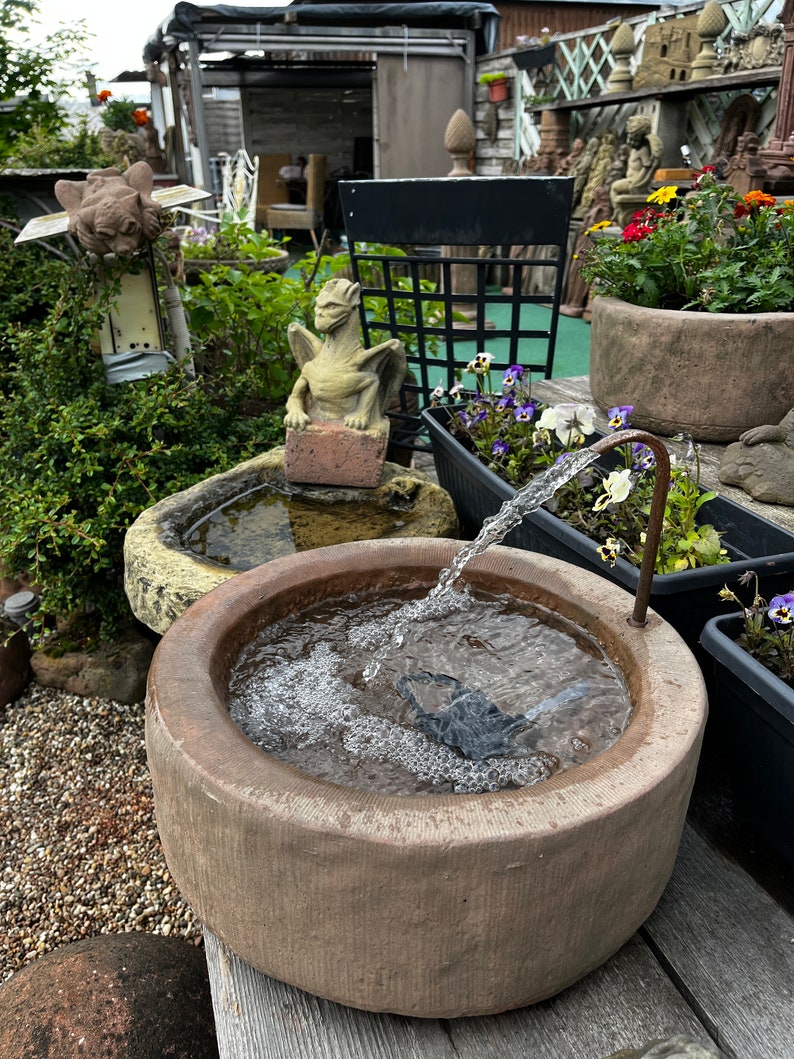May include: A stone fountain with a small dragon statue and a water feature. The fountain is made of brown stone and has a round basin. Water flows from a spout into the basin, creating a small waterfall effect.