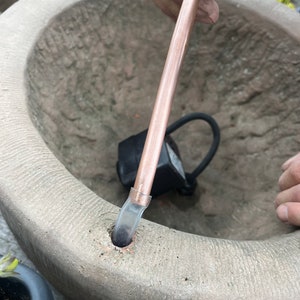 Peut inclure: Un tube en cuivre est ins&eacute;r&eacute; dans une fontaine en pierre. La fontaine a un bassin circulaire avec une surface textur&eacute;e. Une pompe et un tuyau noirs sont visibles &agrave; l'int&eacute;rieur de la fontaine. Le tube est tenu par une personne.