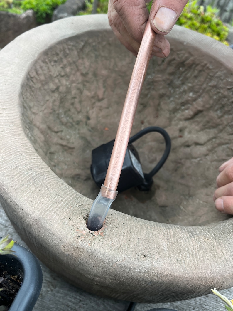 Peut inclure: Un tube en cuivre est ins&eacute;r&eacute; dans une fontaine en pierre. La fontaine a un bassin circulaire avec une surface textur&eacute;e. Une pompe et un tuyau noirs sont visibles &agrave; l'int&eacute;rieur de la fontaine. Le tube est tenu par une personne.