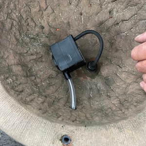 Peut inclure: Une pompe &agrave; eau submersible noire avec un tube en plastique transparent et un tuyau noir incurv&eacute; est plac&eacute;e &agrave; l'int&eacute;rieur d'un grand pot en pierre beige textur&eacute;. Le pot a un aspect rugueux et vieilli.