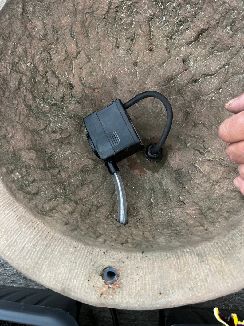 Peut inclure: Une pompe &agrave; eau submersible noire avec un tube en plastique transparent et un tuyau noir incurv&eacute; est plac&eacute;e &agrave; l'int&eacute;rieur d'un grand pot en pierre beige textur&eacute;. Le pot a un aspect rugueux et vieilli.