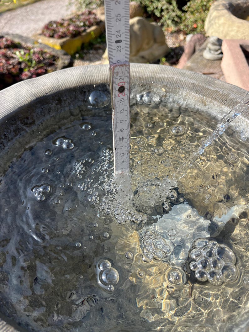 Peut inclure: Un abreuvoir en pierre rempli d'eau, avec une r&egrave;gle indiquant une profondeur d'eau d'environ 19 cm. Des bulles sont visibles dans l'eau, et un filet d'eau coule dans le bassin. L'abreuvoir est &agrave; l'ext&eacute;rieur.