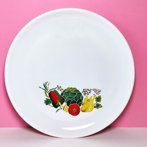 Può includere: Piatto in ceramica bianca con un carciofo verde, peperoni rossi, banane gialle ed erbe verdi stampate su di esso.