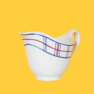Może przedstawiać: Biały dzbanek ceramiczny z niebiesko-czerwonym wzorem w kratkę. Dzbanek ma zakrzywiony uchwyt i zaokrągloną podstawę.