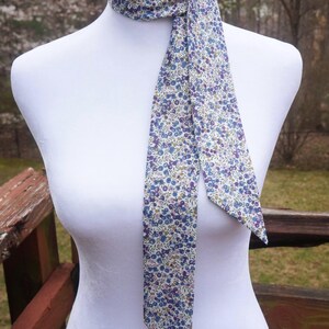 May include: A floral patterned neck scarf in shades of blue, purple, and green on a cream background. The scarf is tied around a mannequin's neck, with the ends hanging down. The fabric has a delicate, small-scale floral print.