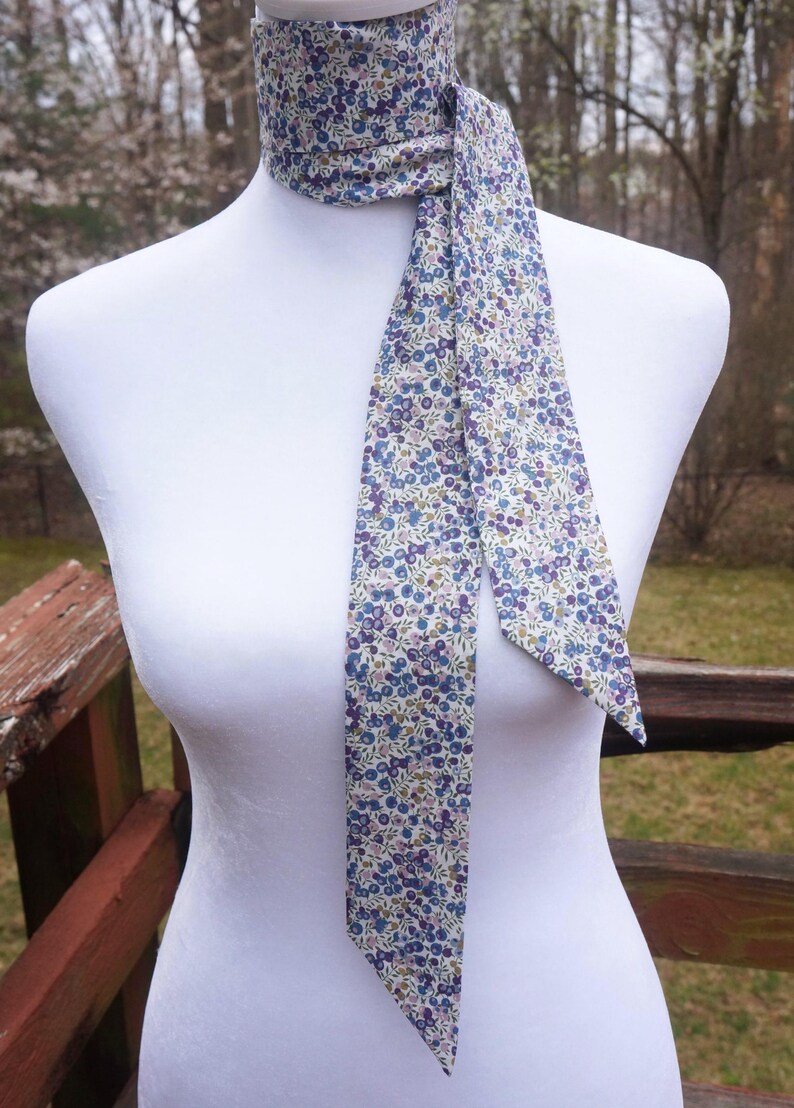 May include: A floral patterned neck scarf in shades of blue, purple, and green on a cream background. The scarf is tied around a mannequin's neck, with the ends hanging down. The fabric has a delicate, small-scale floral print.