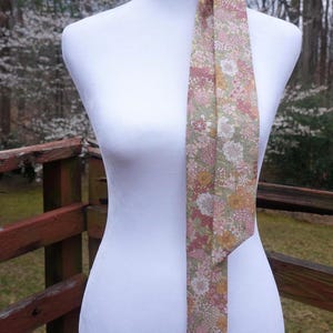 May include: A floral patterned neck scarf in shades of pink, yellow, and white on a light green background. The scarf is draped over a white mannequin, with a wooden railing and trees in the background.
