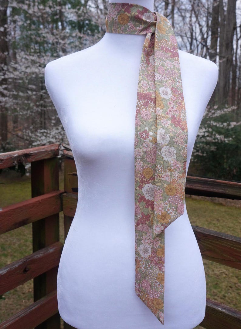 May include: A floral patterned neck scarf in shades of pink, yellow, and white on a light green background. The scarf is draped over a white mannequin, with a wooden railing and trees in the background.