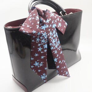 May include: A glossy black handbag with a burgundy interior and handle. A burgundy scarf with a blue floral pattern is tied to the handle. The bag has a rectangular shape and a shiny finish.