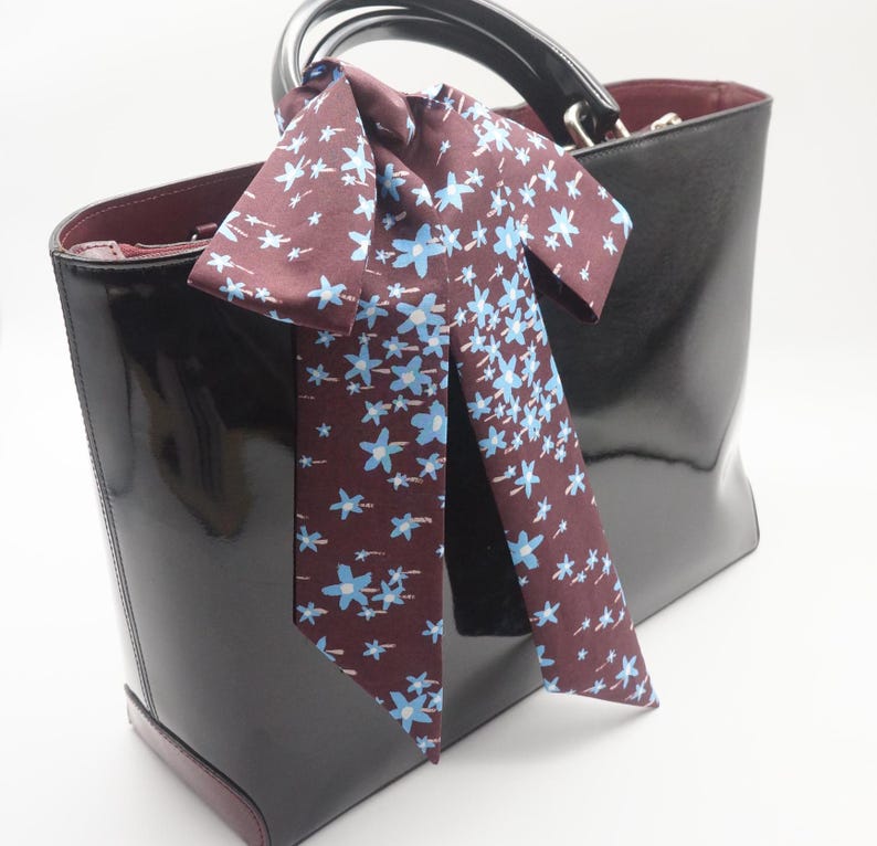 May include: A glossy black handbag with a burgundy interior and handle. A burgundy scarf with a blue floral pattern is tied to the handle. The bag has a rectangular shape and a shiny finish.