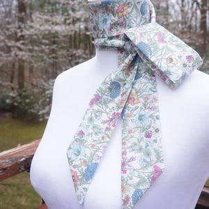 May include: A floral patterned neck scarf with a matching pocket square. The scarf and pocket square feature a white background with a colorful floral design in shades of blue, pink, orange, and green. The scarf is draped around a white mannequin.