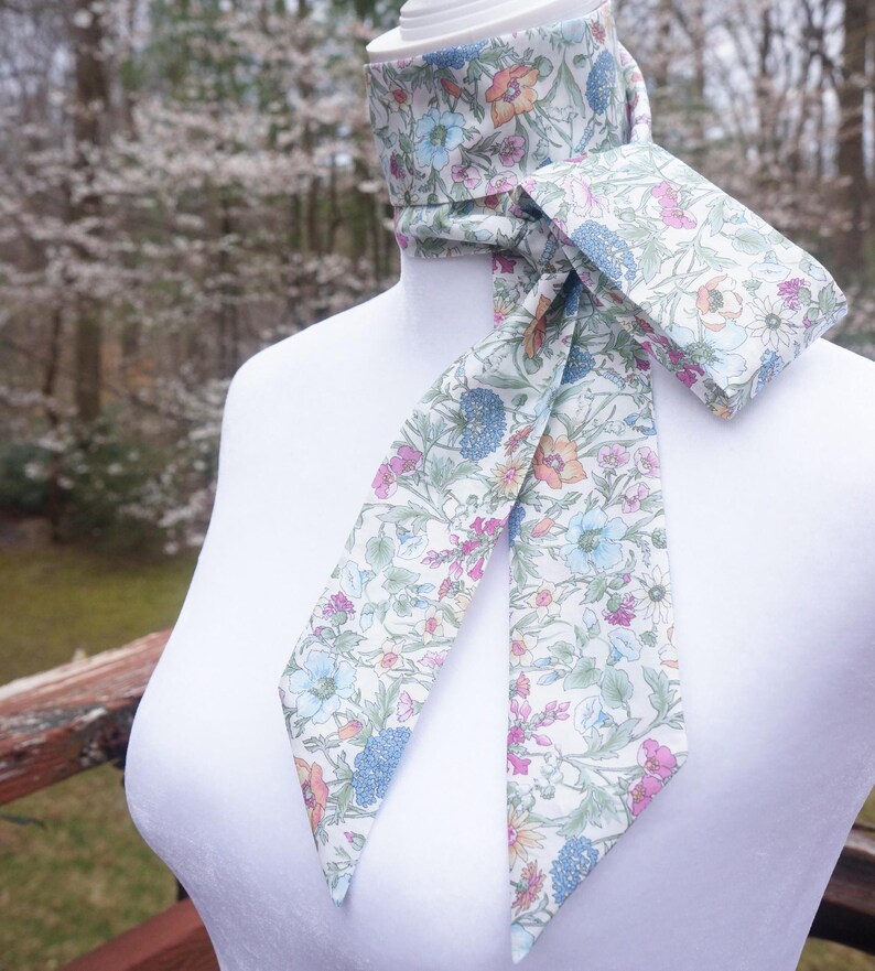 May include: A floral patterned neck scarf with a matching pocket square. The scarf and pocket square feature a white background with a colorful floral design in shades of blue, pink, orange, and green. The scarf is draped around a white mannequin.
